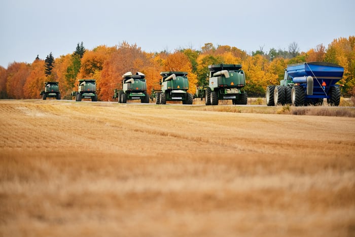 A line of large farming vehicles rolls down the side of a field in early Autumn. The field is a light brown color.