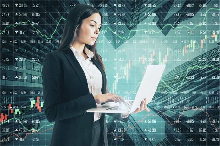 A person standing typing on a laptop with stock market quotes in the background.