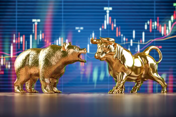 A golden bear growls at a golden bull, both of which stand in front of a stock price chart.