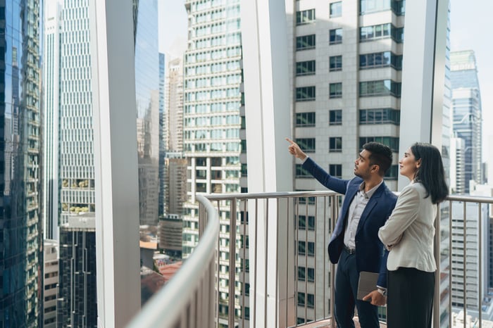 Two people on balcony, looking up at city buildings.