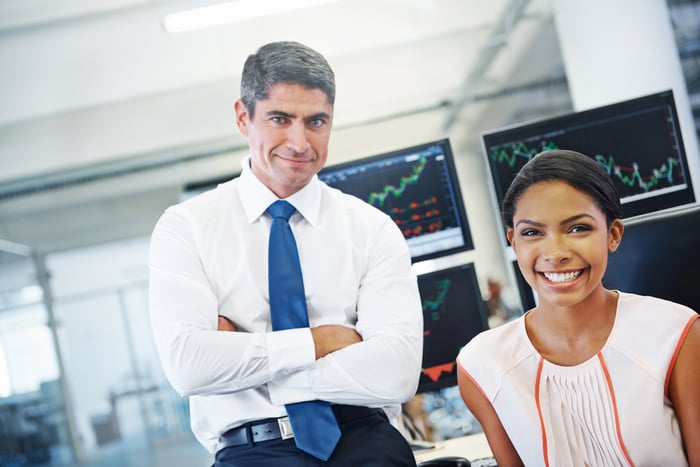 Investment analysts are smiling while stock charts can be seen on terminals behind them.