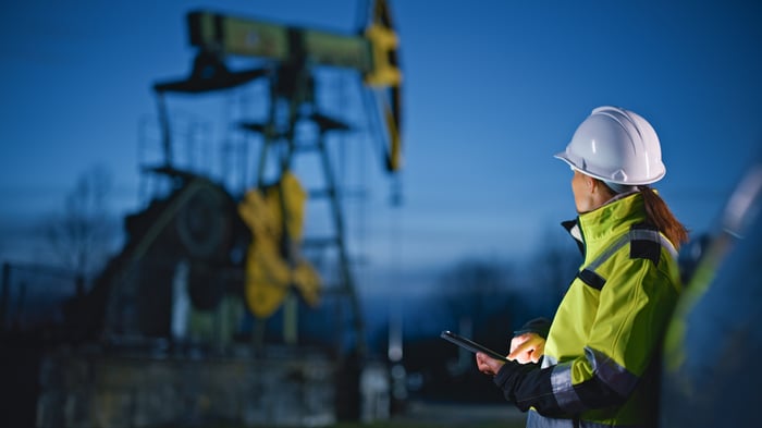 An oilfield worker holds a tablet while looking at a pumpjack in the distance.