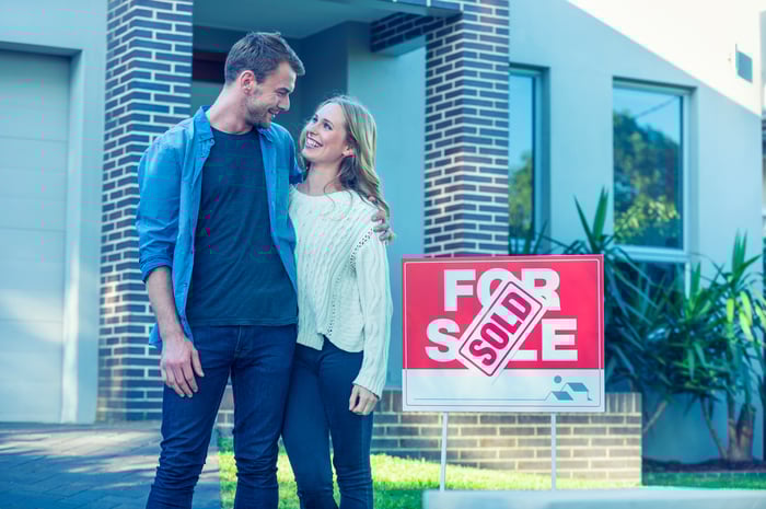 Two people are smiling while standing near a sold sign in front of a home.