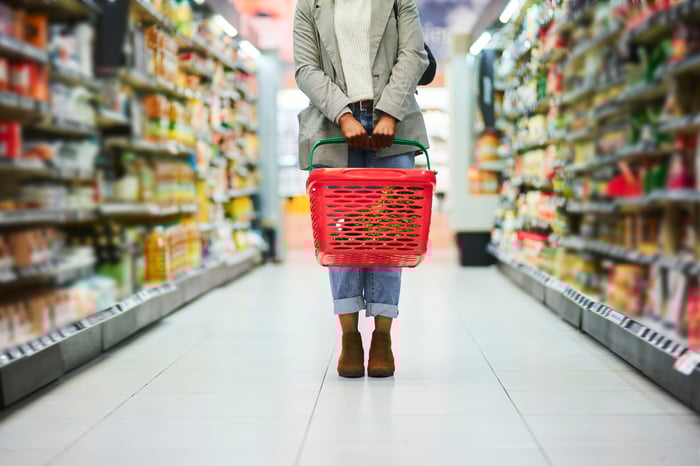 A person carries a grocery basket through a supermarket aisle. 