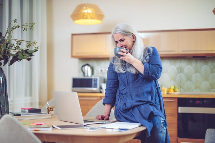 Person standing at a desk, looking down on a laptop as they sip coffee.