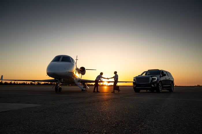 Two people between a plane and a car shake hands at sunset on an airport tarmac.