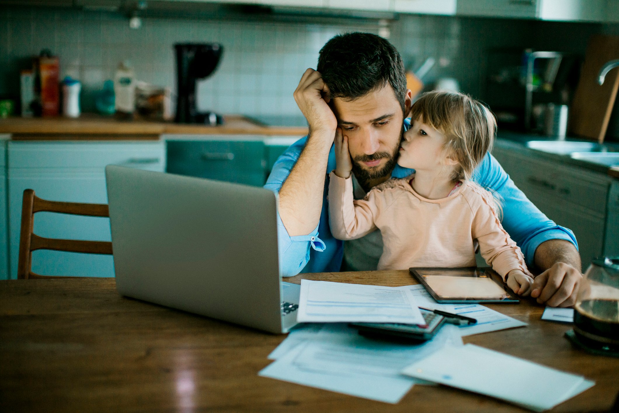 Stressed person looking at laptop and documents with young child sitting on lap kissing their face