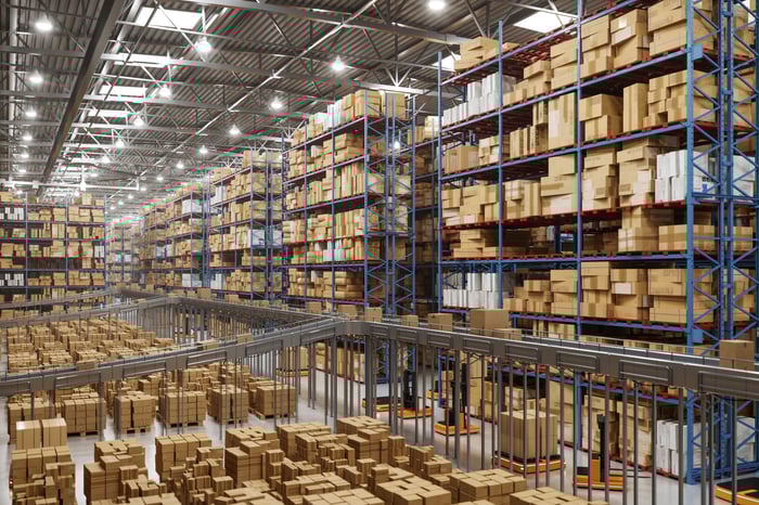 A vast, high-ceilinged warehouse features rows of tall blue shelving units stacked high with cardboard boxes and automated carts moving along the floor.