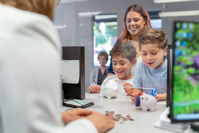 A parent and two young kids in a bank counting coins from their piggy banks.
