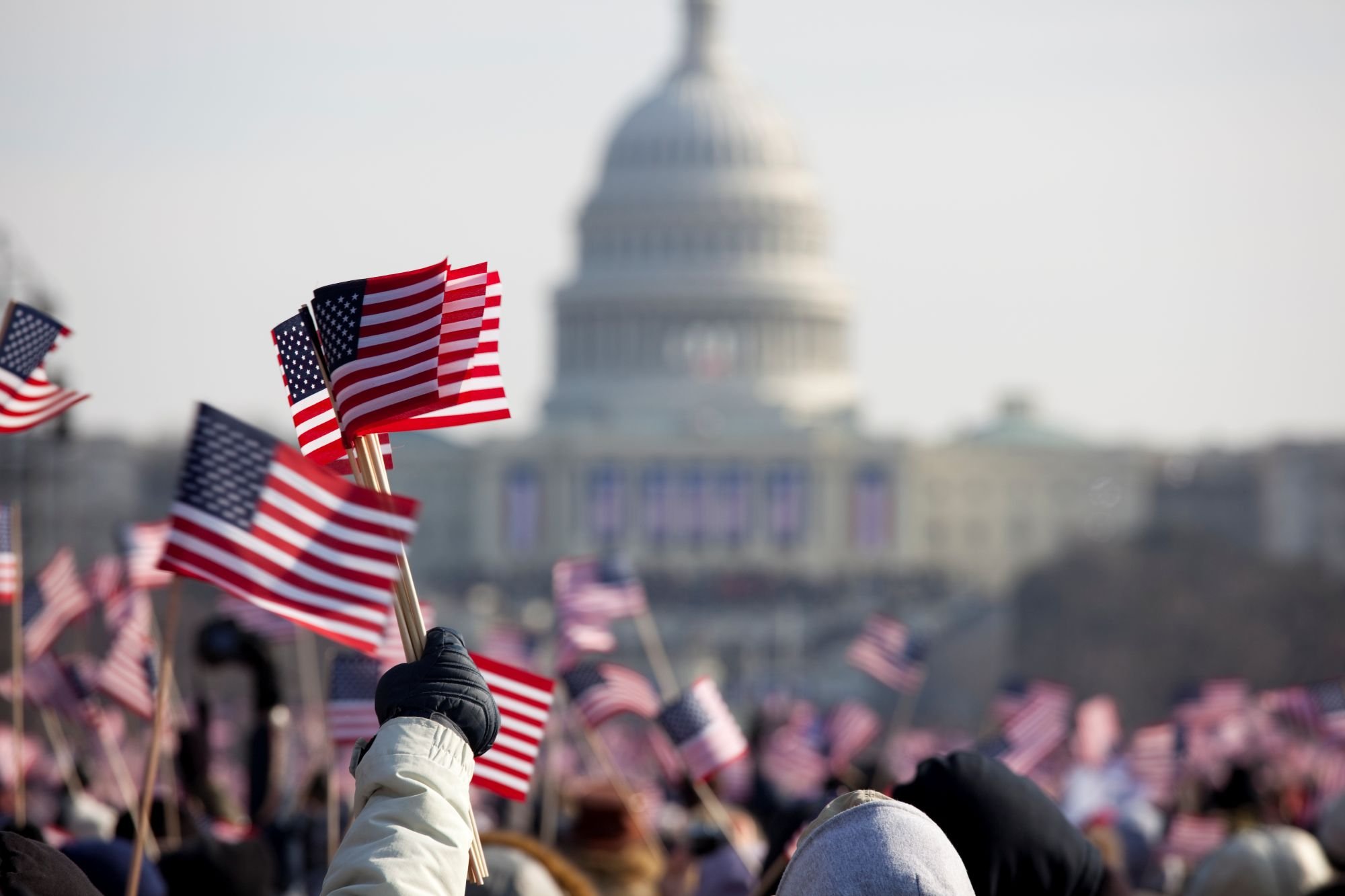 A gloved hand holds several small American flags aloft in the foreground of a large crowd gathered before the blurred silhouette of the United States Capitol building.