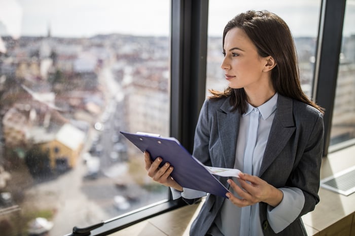 An investor stands in an office while looking out a window and holding a clipboard with some documents.