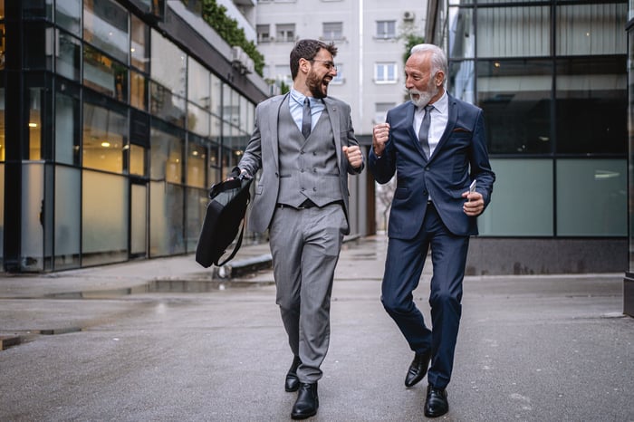 Two happy men dressed in business suits walking together on an urban street and bumping their fists.