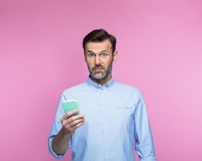 Investor in disbelief wearing a button-down shirt while holding a smartphone.