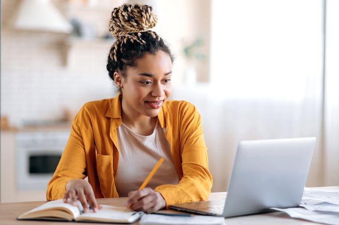 A person, smiling, wriiting down notes on a pad, looking at their laptop. 