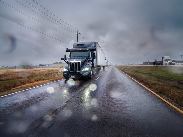 Long-haul truck in rainy weather on a long stretch of highway through farmland.