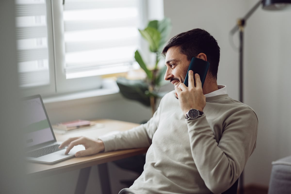 An investor smiles as they sit at a desk, working on a computer and talking on the phone.