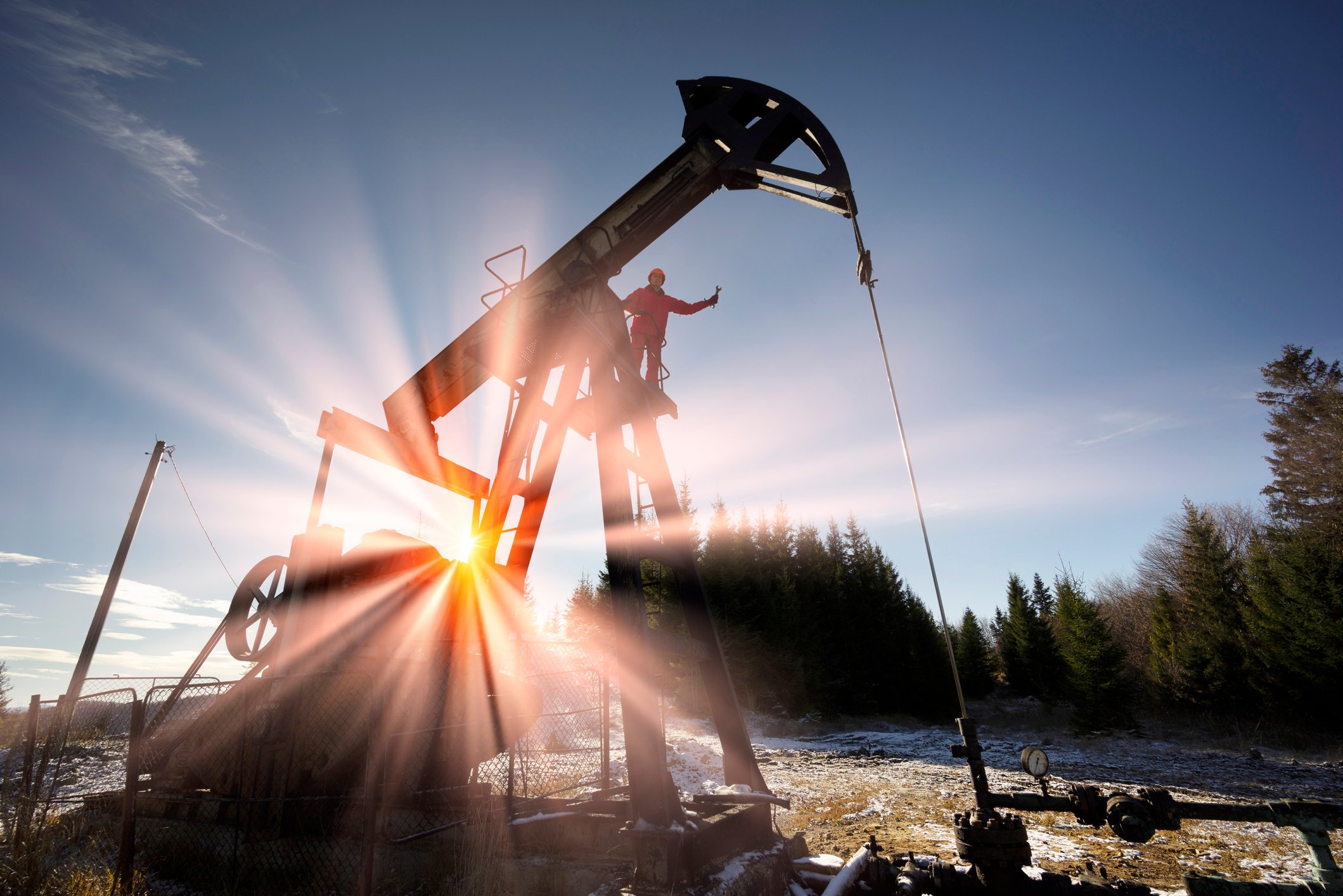 A person standing on an oil well with the sun shining in the background.