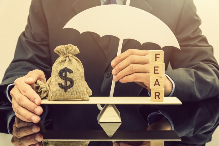 A person balancing a scale with a moneybag on one side and blocks spelling "fear" on the other, with an umbrella in the center.