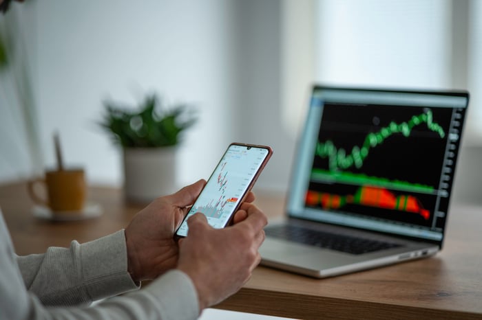 A person trading on a mobile App with a laptop displaying stock price charts placed in front.