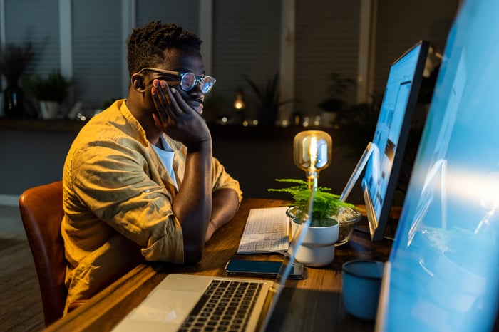 An investor sits in front of two computers and a laptop, working late at night.
