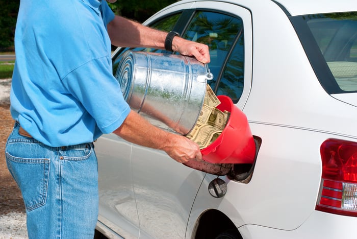 A person dumping a bucket of cash into the gas tank of a car.
