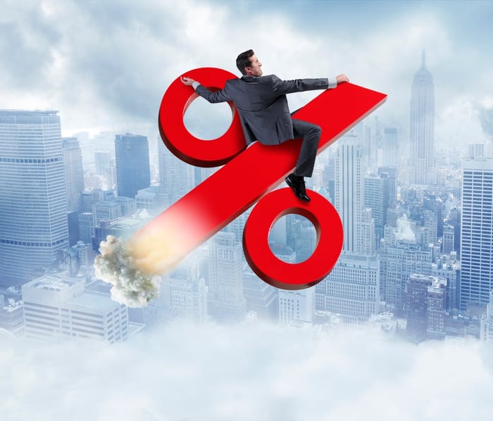 A man in a business suit riding a giant red interest rate sign into the sky.