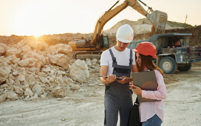 Two workers in hard hats reviewing notes at a mine with an excavator loading rocks in a truck in the background.