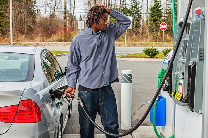 A person puts their hand to their head in surprise while filling up a silver car at a gas station.