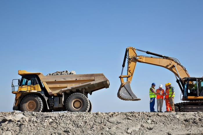 A dump truck and an excavator are pictured against a blue sky. Four construction workers stand in a group between the machines.