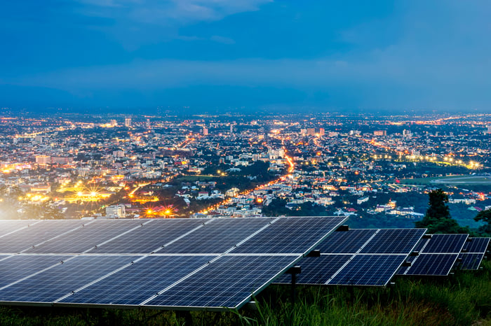 An array of solar panels against a background of a large city at dusk.