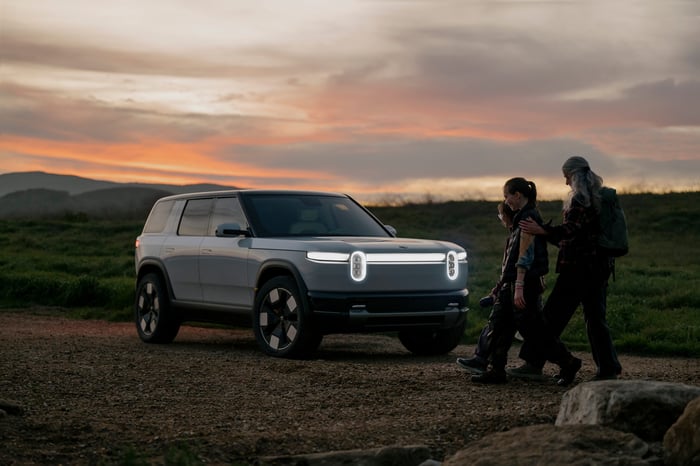 Two people walk next to a Rivian R2 vehicle with a orange-grey sky and mountains in the background