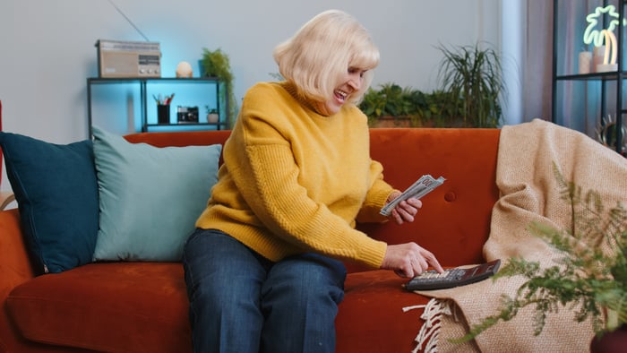 An individual - presumably retired- punches numbers into a calculator while sitting on a sofa.