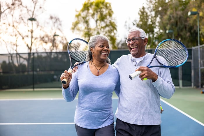 Older couple, laughing together as they walk off a tennis court. 