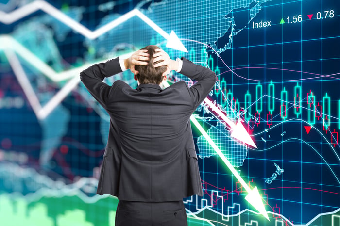 A person in a gray business suit looks away from the camera at a large chart of downward arrows with their hands on their head.