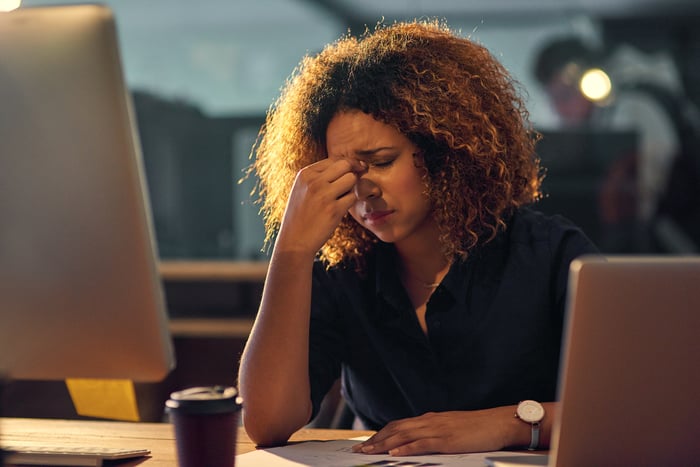 An investor touches the bridge of nose in frustration while sitting at a desk in front of two computers.