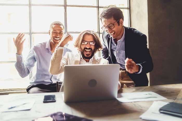 Three investors gather around a laptop and cheer what they're seeing. 
