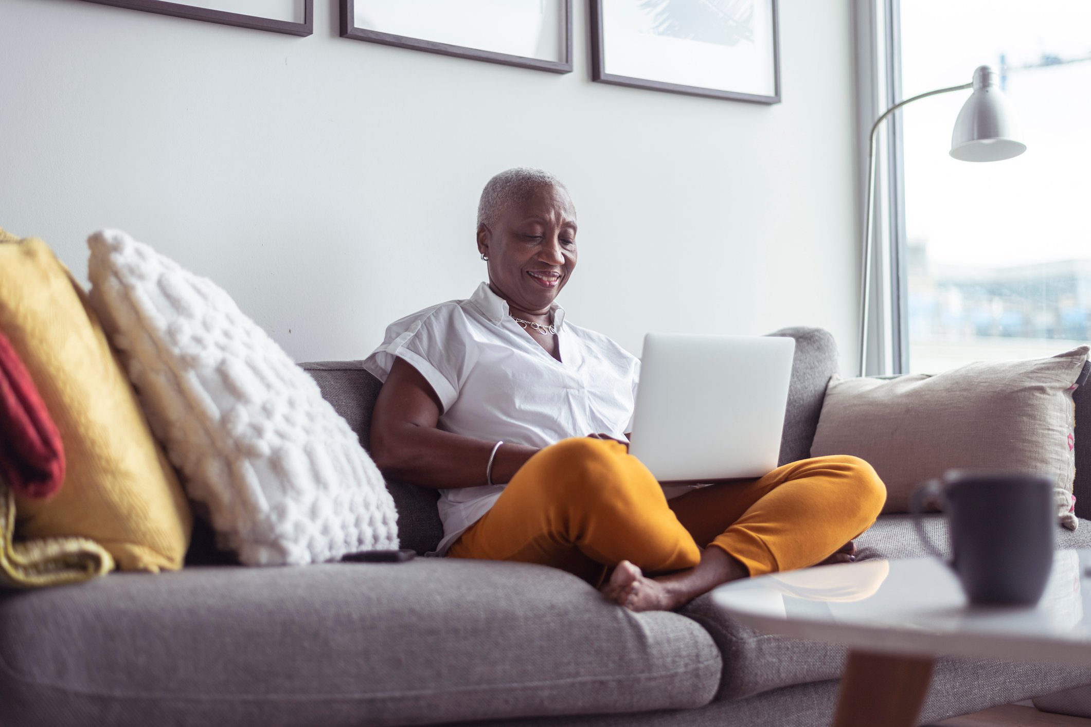 Senior smiling and sitting cross legged on couch using laptop
