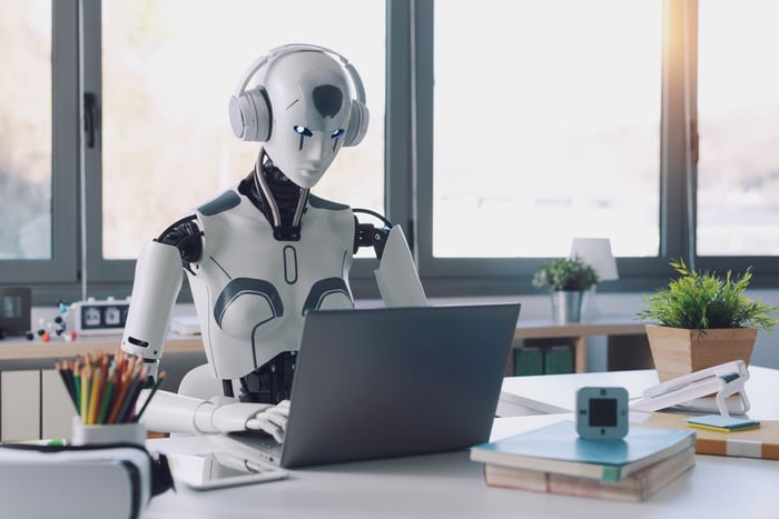 Robot sitting at a desk, appearing to work on a laptop.