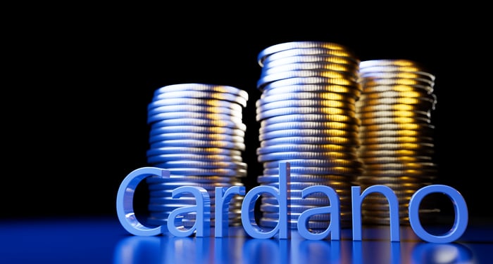 A pile of coins lie stacked behind big letters that spell "Cardano."