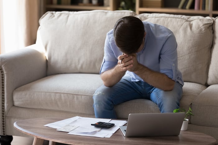 Person sitting on sofa with head in hands, in front of table with a laptop and papers. 