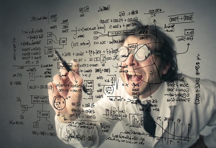 A person in thick glasses and a lab coat writes a complicated equation on a transparent board.