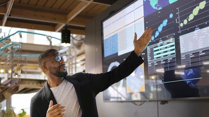 A person pointing up at charts on screens in an office setting. 