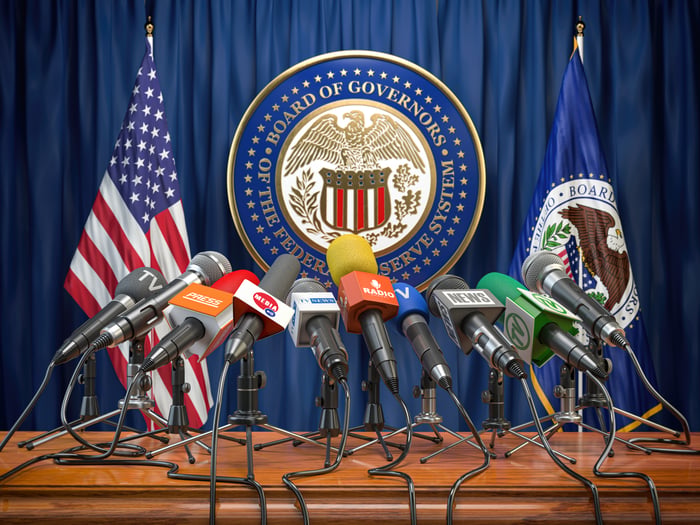 A bunch of microphones at an empty podium with the symbol of the Federal Reserve Board in the background. 