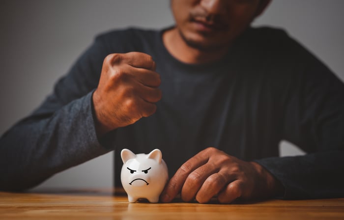 A person holds their fist over a frowning piggy bank.