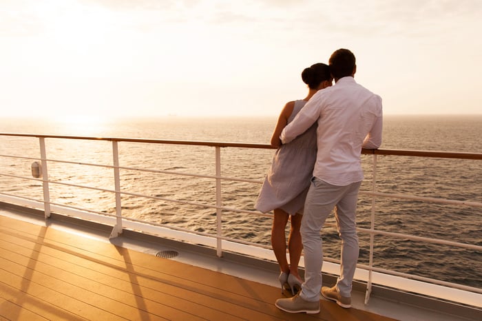 A couple on a cruise ship looks out at the ocean and the setting sun.
