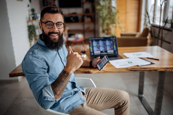 An investor gives a thumbs up and expresses approval while sitting at a desk in front of a computer and holding a phone.
