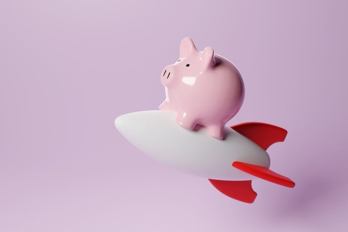 A pink ceramic piggy bank riding a white rocket with red fins against a lavender background.