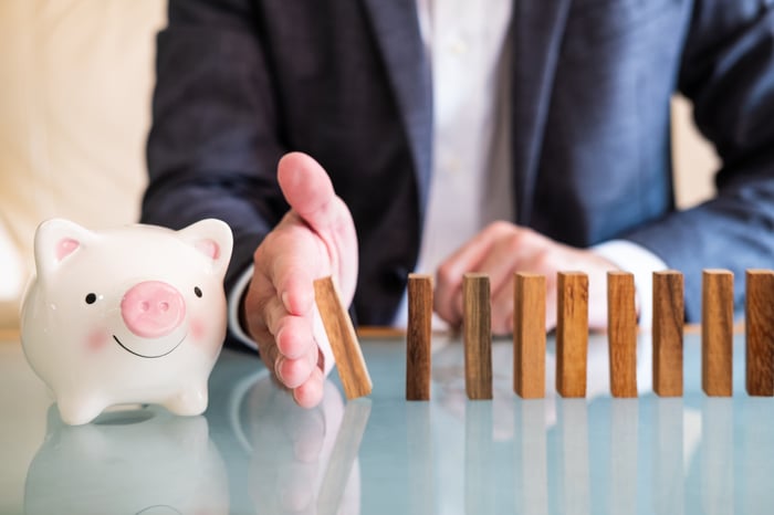 A person stops a line of falling domino tiles with their hand before it touches a smiling piggy bank.
