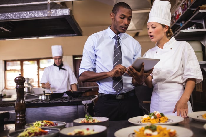 A chef and a manager talking over a tablet computer in a restaurant kitchen.