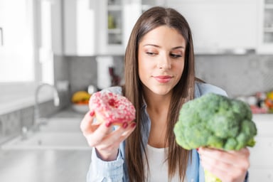 A person in a kitchen decides between eating a doughnut and broccoli.
